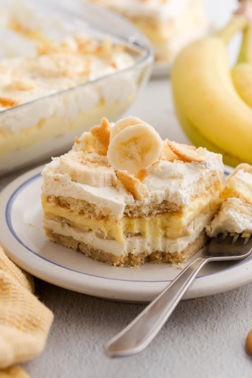 A close-up side-angle shot of a single serving of banana pudding in a clear glass, showing distinct layers of vanilla wafers, banana slices, and creamy pudding topped with a swirl of whipped cream and a whole vanilla wafer