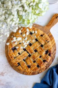 Golden lattice-top blueberry pie cooling on a wire rack with a dark purple bubbling filling visible through the woven crust, surrounded by fresh blueberries on a rustic wooden table