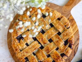 Golden lattice-top blueberry pie cooling on a wire rack with a dark purple bubbling filling visible through the woven crust, surrounded by fresh blueberries on a rustic wooden table