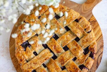 Golden lattice-top blueberry pie cooling on a wire rack with a dark purple bubbling filling visible through the woven crust, surrounded by fresh blueberries on a rustic wooden table