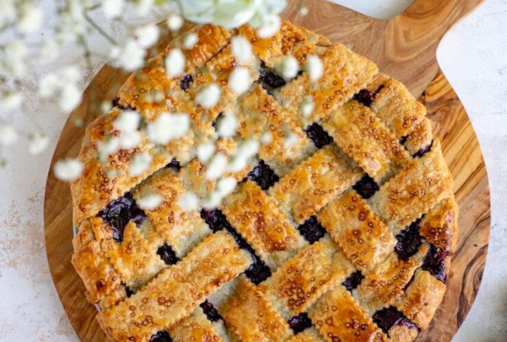Golden lattice-top blueberry pie cooling on a wire rack with a dark purple bubbling filling visible through the woven crust, surrounded by fresh blueberries on a rustic wooden table