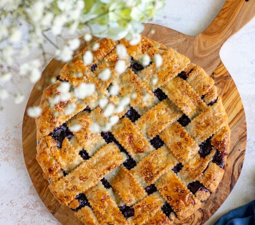 Golden lattice-top blueberry pie cooling on a wire rack with a dark purple bubbling filling visible through the woven crust, surrounded by fresh blueberries on a rustic wooden table