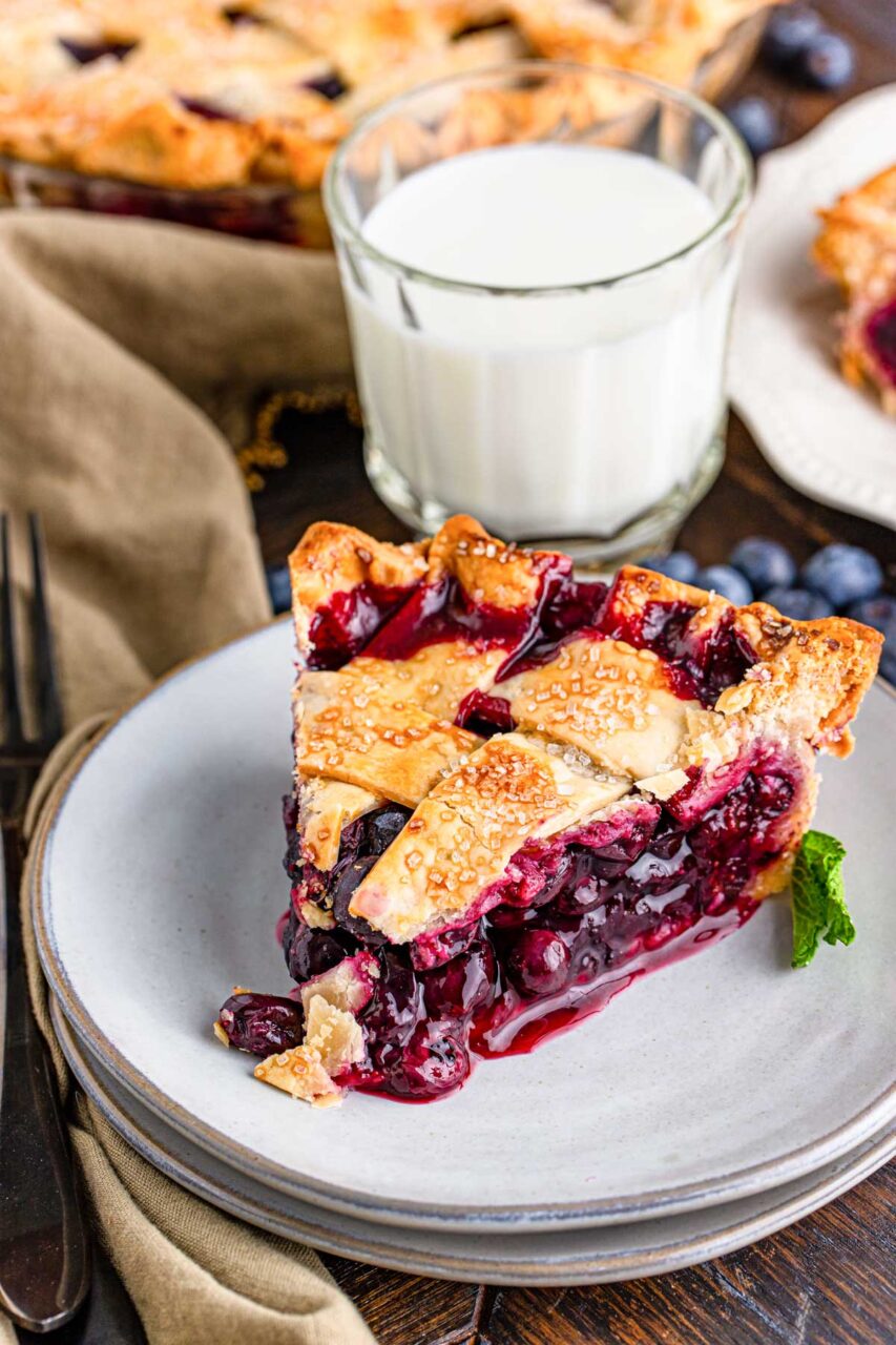 Step-by-step flat lay showing blueberry pie filling ingredients — fresh blueberries, sugar, cornstarch, lemon zest, and butter — alongside strips of rolled pie dough arranged for a lattice top on a floured surface