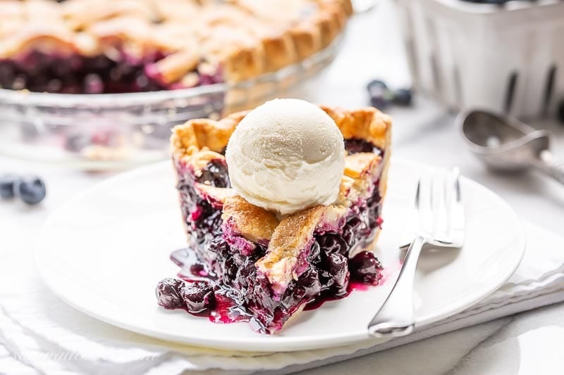 Close-up slice of blueberry pie on a white plate showing the deep purple jammy filling and flaky golden lattice crust layers, topped with a scoop of vanilla ice cream melting at the edges