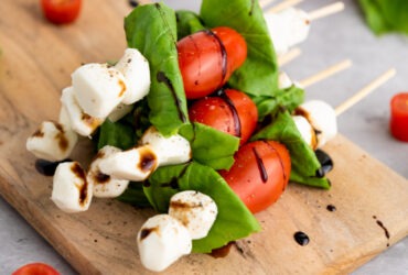 Caprese skewers arranged on a white marble board with balsamic glaze drizzle, fresh basil leaves, and cherry tomatoes