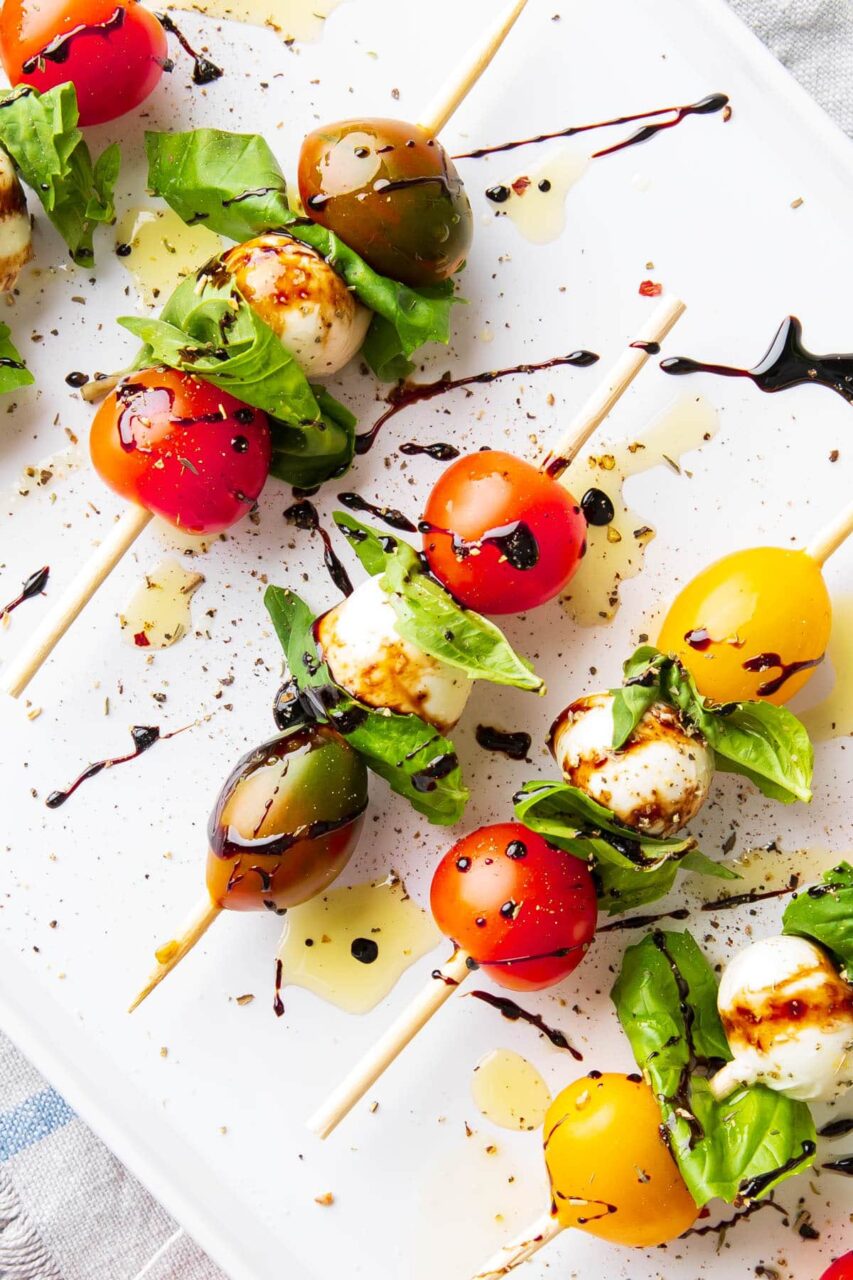 Ingredients for Caprese skewers laid out flat on a wooden cutting board — cherry tomatoes, mozzarella balls, fresh basil, olive oil, and balsamic glaze