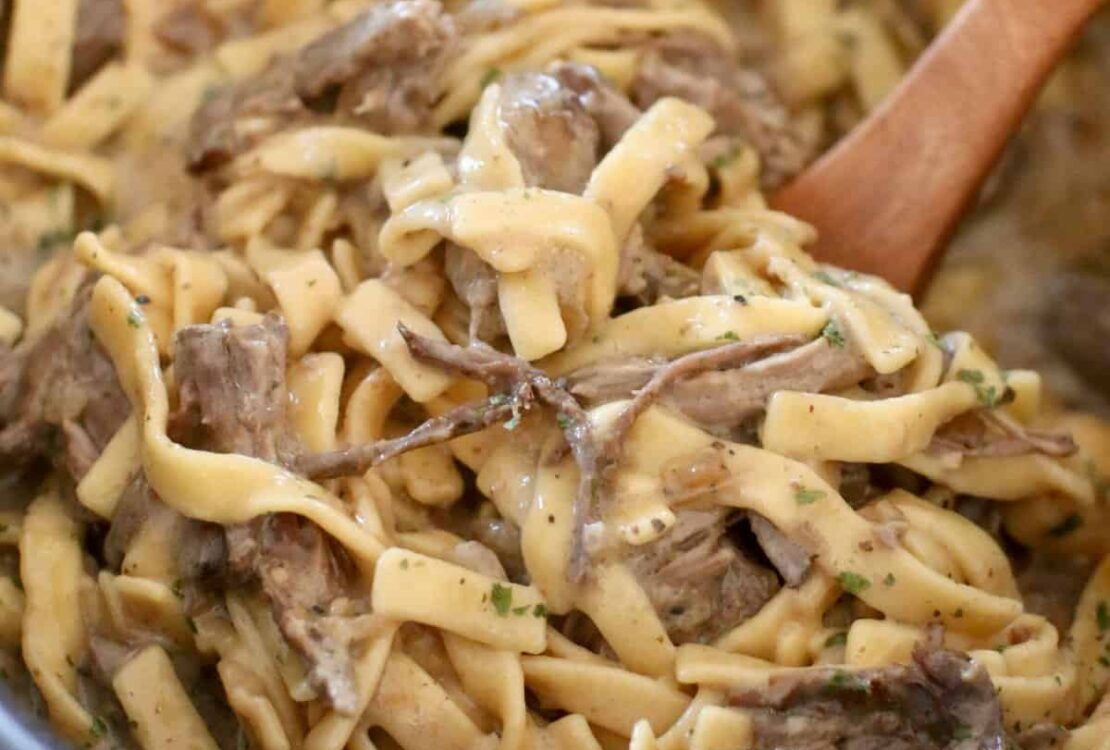 Crockpot beef and noodles served in a wide bowl with tender shredded beef over thick egg noodles coated in rich brown gravy, garnished with fresh parsley