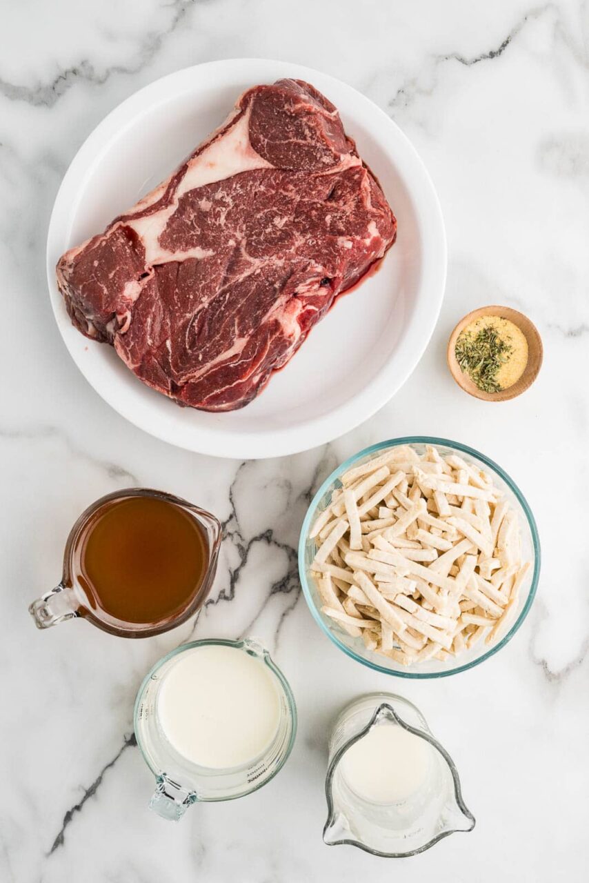 Overhead flat lay of crockpot beef and noodles ingredients including chuck roast, egg noodles, beef broth, Worcestershire sauce, onion, garlic, and cornstarch arranged on a wooden surface
