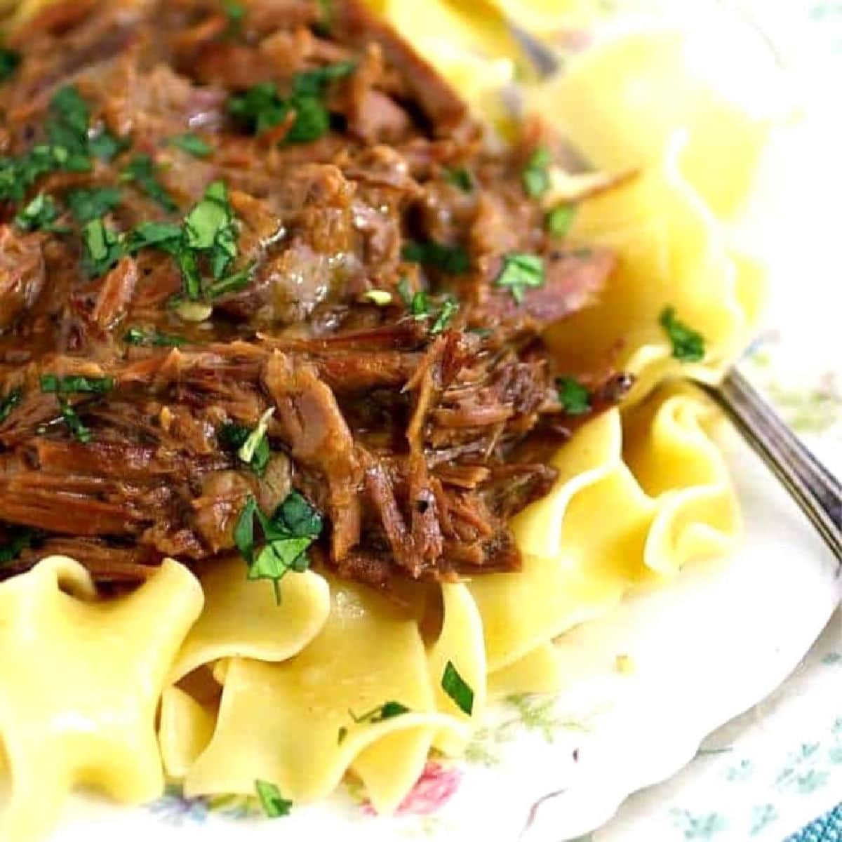 Close-up of a fork lifting tender shredded beef from a bowl of crockpot beef and noodles, showing the glossy rich brown gravy coating the egg noodles