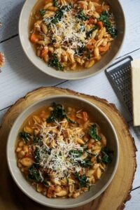 A rustic white bowl filled with Crockpot Pasta e Fagioli — thick tomato broth with ditalini pasta, kidney beans, cannellini beans, and ground beef, garnished with freshly grated Parmesan and chopped fresh parsley, photographed on a wooden farmhouse table with crusty bread on the side