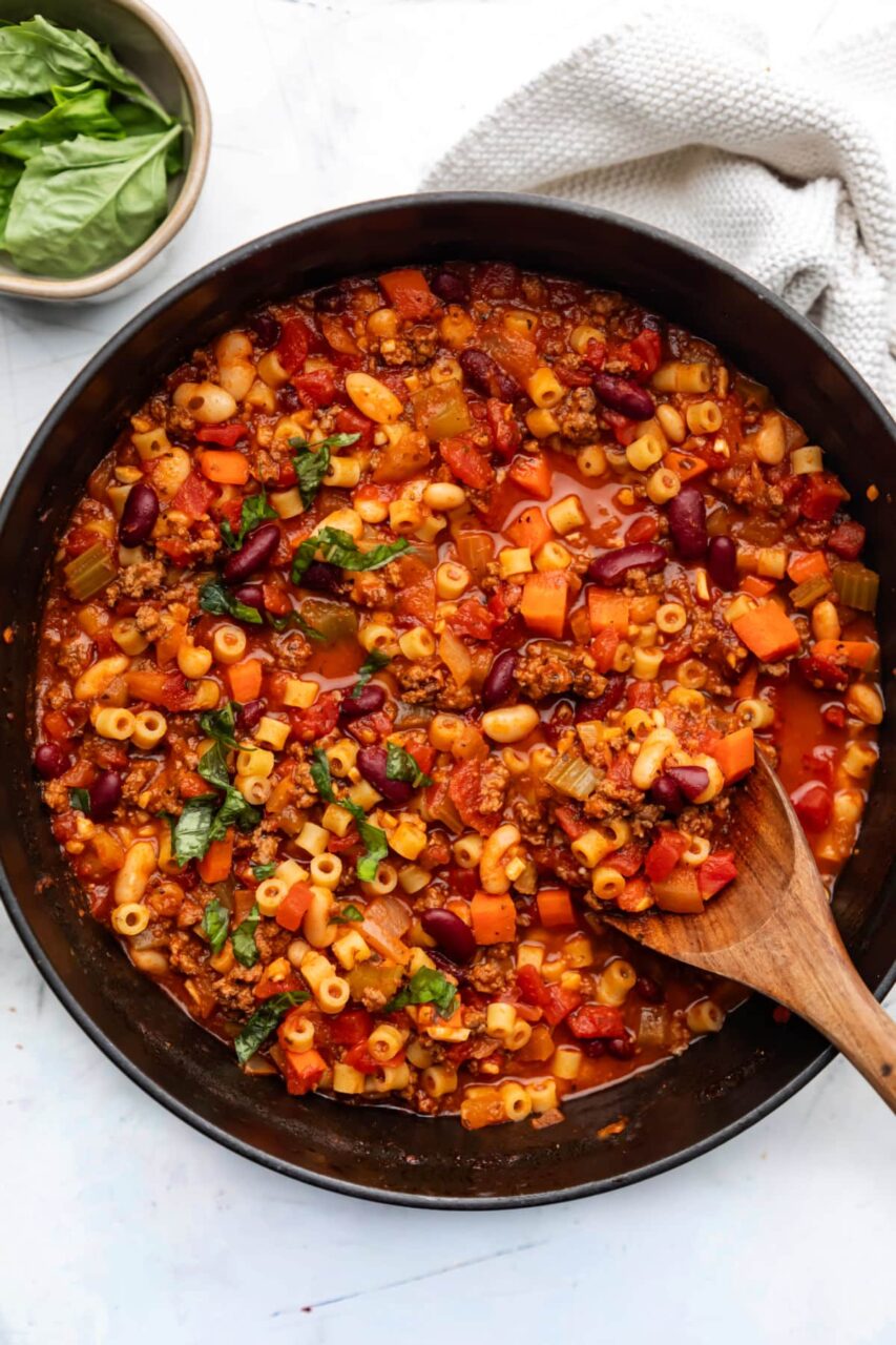 A flat lay of the Crockpot Pasta e Fagioli ingredients arranged on a white surface — cans of kidney beans, cannellini beans, diced and crushed tomatoes, raw ditalini pasta, raw ground beef, chicken broth carton, fresh garlic, onion, celery, carrots, and a small bowl of Italian seasoning