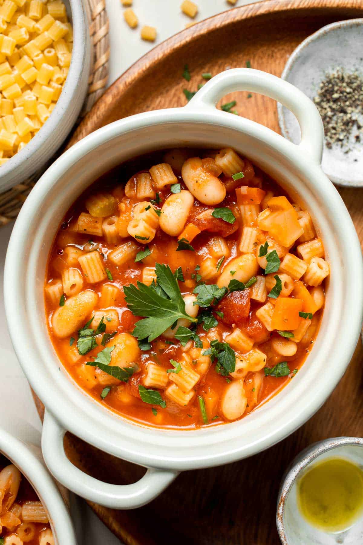 A close-up overhead shot of a bowl of Crockpot Pasta e Fagioli with a spoon lifting a scoop of ditalini pasta, kidney beans, and rich red tomato broth, topped with freshly shaved Parmesan cheese and a drizzle of olive oil, with a piece of crusty Italian bread in the background