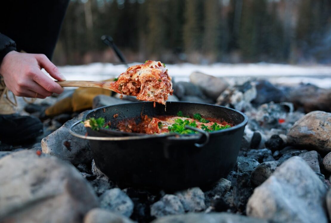 Dutch Oven Lasagna bubbling with melted cheese and sausage sauce, served beside a campfire on a rustic wooden board