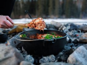 Dutch Oven Lasagna bubbling with melted cheese and sausage sauce, served beside a campfire on a rustic wooden board