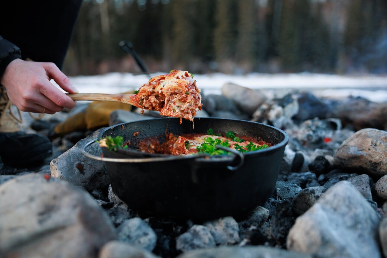 Dutch Oven Lasagna bubbling with melted cheese and sausage sauce, served beside a campfire on a rustic wooden board