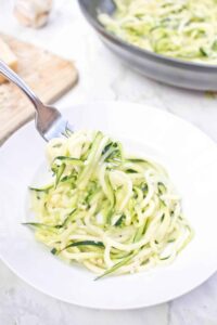 Garlic Parmesan Zoodles beautifully plated in a white pasta bowl, topped with freshly grated Parmesan, cracked black pepper, and fresh basil, shot overhead on a rustic wood surface