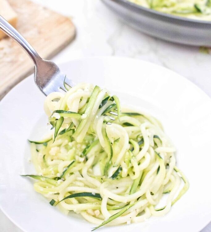 Garlic Parmesan Zoodles beautifully plated in a white pasta bowl, topped with freshly grated Parmesan, cracked black pepper, and fresh basil, shot overhead on a rustic wood surface