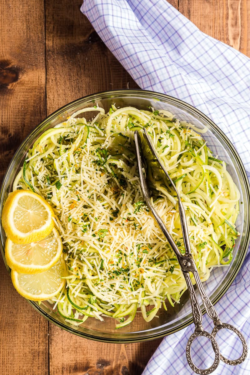 Flat lay of garlic Parmesan zoodles ingredients including spiralized zucchini noodles in a colander, minced garlic, a block of Parmesan being grated, butter, olive oil, and lemon on a marble surface