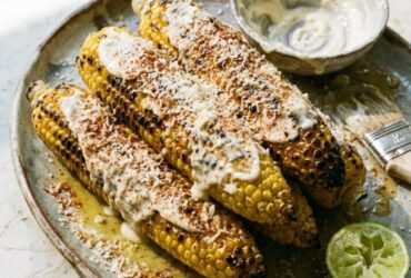 Grilled corn on the cob elote style with charred kernels, Parmesan, chili butter, and lime wedges on a rustic wooden board