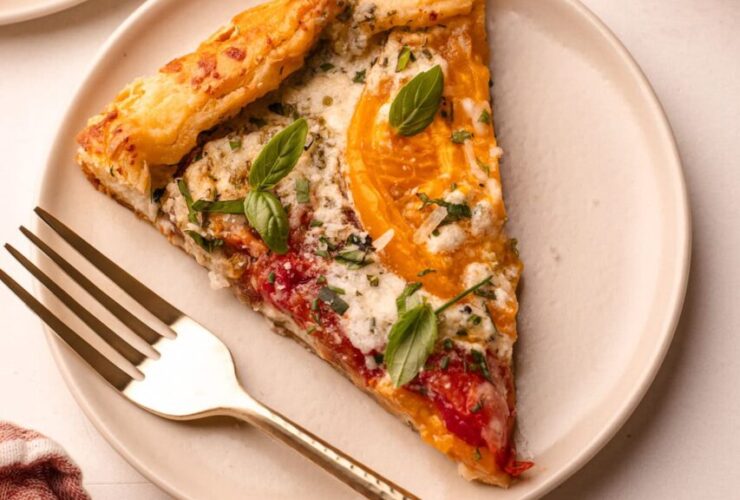 A beautifully plated heirloom tomato galette on a wooden serving board, golden flaky crust folded over vibrant red and yellow heirloom tomato slices, garnished with fresh basil leaves and a drizzle of olive oil