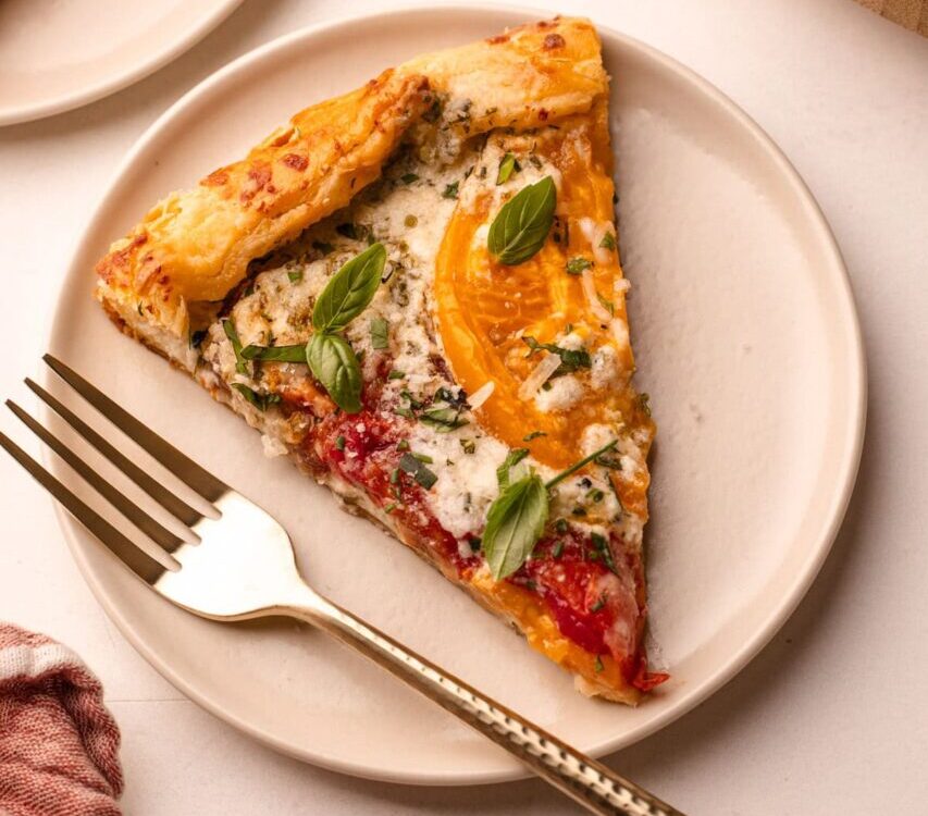 A beautifully plated heirloom tomato galette on a wooden serving board, golden flaky crust folded over vibrant red and yellow heirloom tomato slices, garnished with fresh basil leaves and a drizzle of olive oil