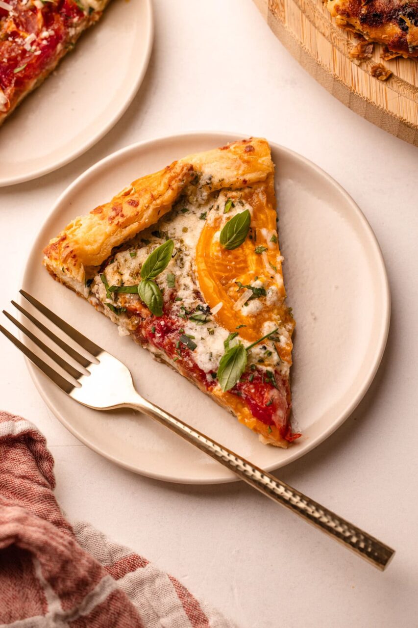 A beautifully plated heirloom tomato galette on a wooden serving board, golden flaky crust folded over vibrant red and yellow heirloom tomato slices, garnished with fresh basil leaves and a drizzle of olive oil