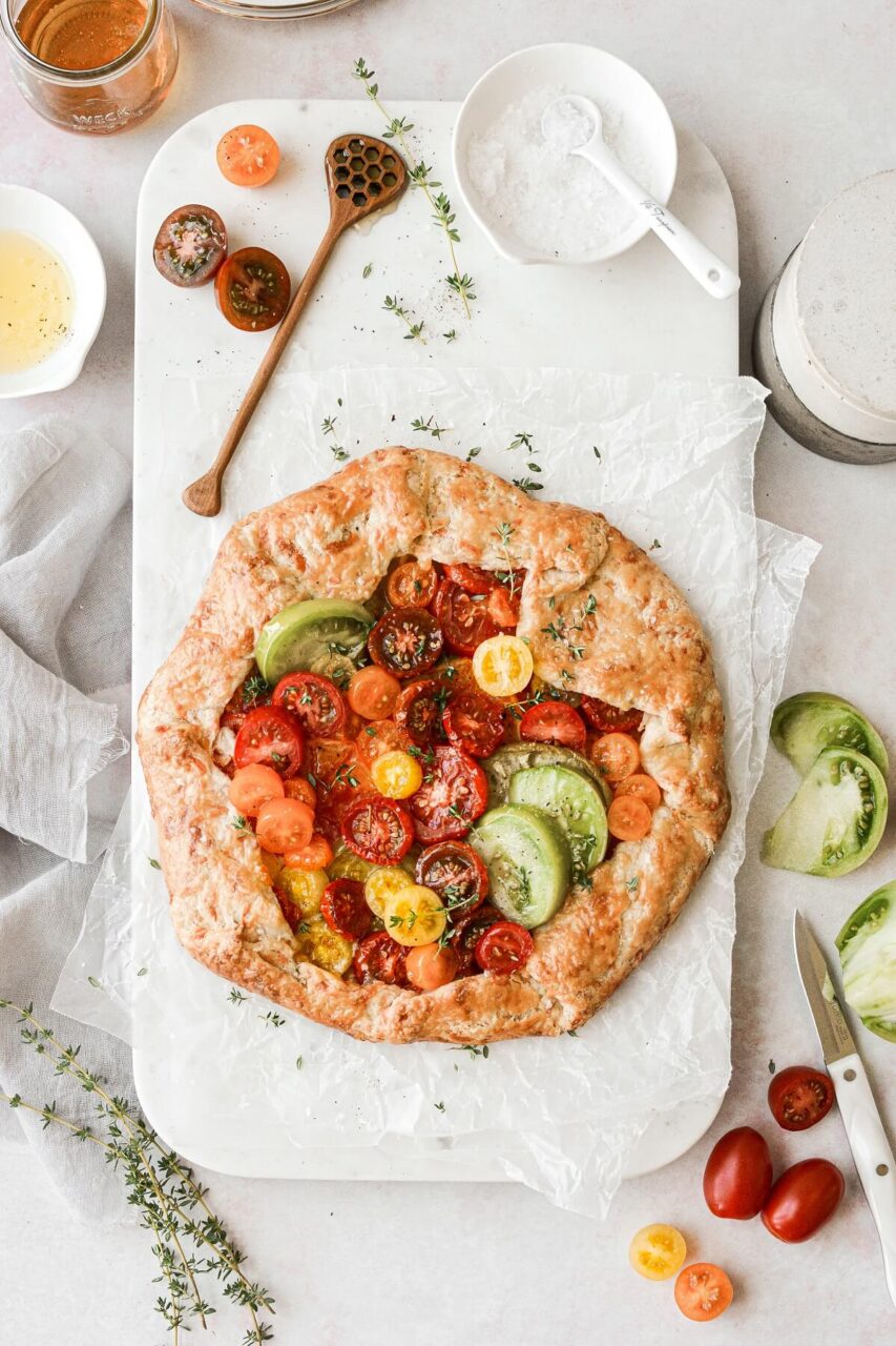 Overhead flat-lay of heirloom tomato galette ingredients on a marble surface — sliced colorful tomatoes draining on paper towels, ricotta in a bowl, fresh basil, garlic cloves, rolled-out pie dough on a floured surface, and a block of Parmesan