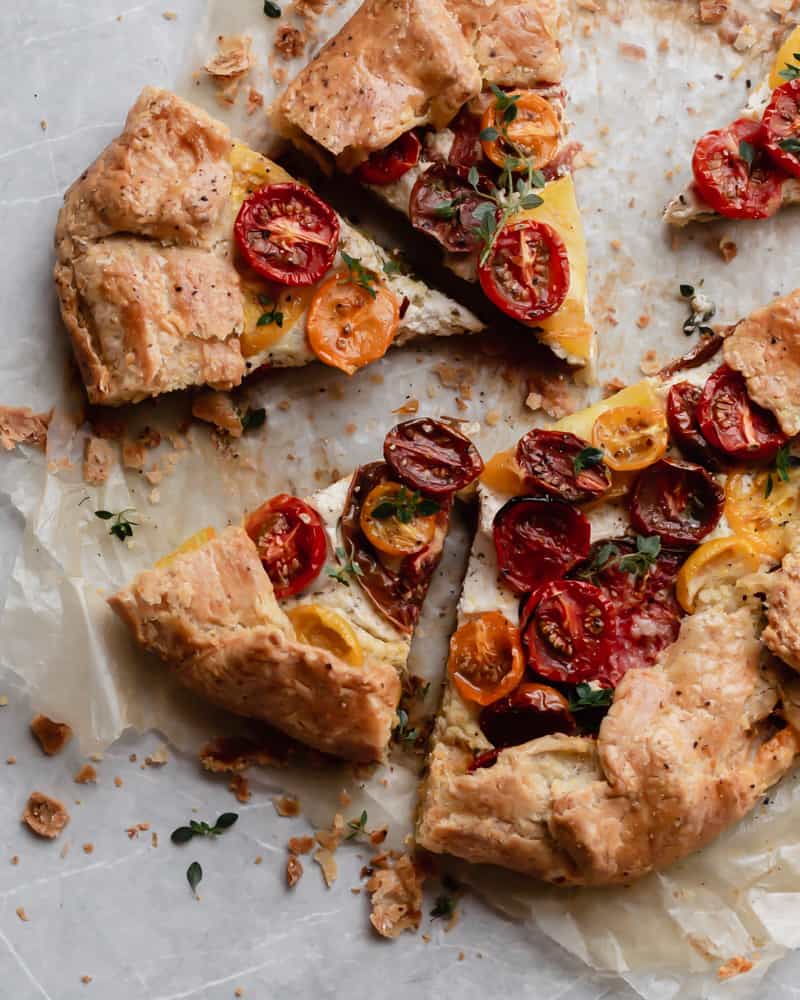 Close-up of a slice of heirloom tomato galette being lifted from the tart, showing the golden flaky crust layers, creamy ricotta base, and jewel-toned roasted tomato slices topped with a fresh basil leaf