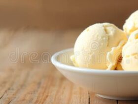 Homemade vanilla ice cream scooped into a white ceramic bowl, golden and creamy with vanilla bean specks, garnished with a fresh vanilla pod on a rustic wooden table in soft summer light