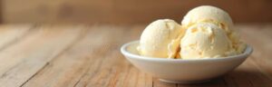 Homemade vanilla ice cream scooped into a white ceramic bowl, golden and creamy with vanilla bean specks, garnished with a fresh vanilla pod on a rustic wooden table in soft summer light