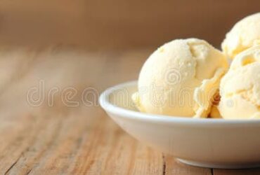 Homemade vanilla ice cream scooped into a white ceramic bowl, golden and creamy with vanilla bean specks, garnished with a fresh vanilla pod on a rustic wooden table in soft summer light
