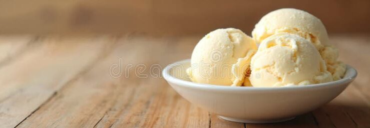 Homemade vanilla ice cream scooped into a white ceramic bowl, golden and creamy with vanilla bean specks, garnished with a fresh vanilla pod on a rustic wooden table in soft summer light