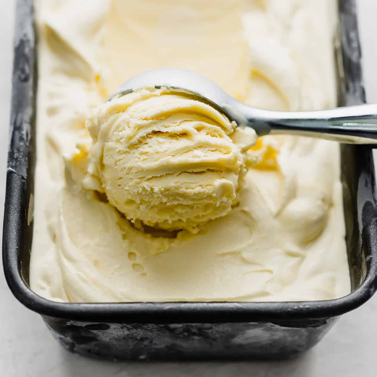 Step-by-step flat lay showing vanilla ice cream ingredients — heavy cream, egg yolks, sugar, vanilla bean split open, and whole milk on a white marble surface with a saucepan and whisk