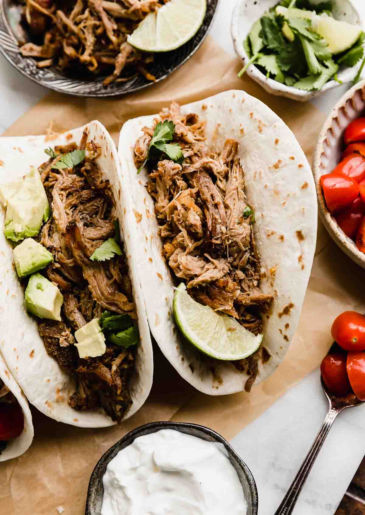 Overhead flat lay of Instant Pot carnitas ingredients — cubed pork shoulder pieces, whole garlic cloves, halved orange and lime, small bowls of cumin, chili powder, smoked paprika, and dried oregano on a rustic wooden surface