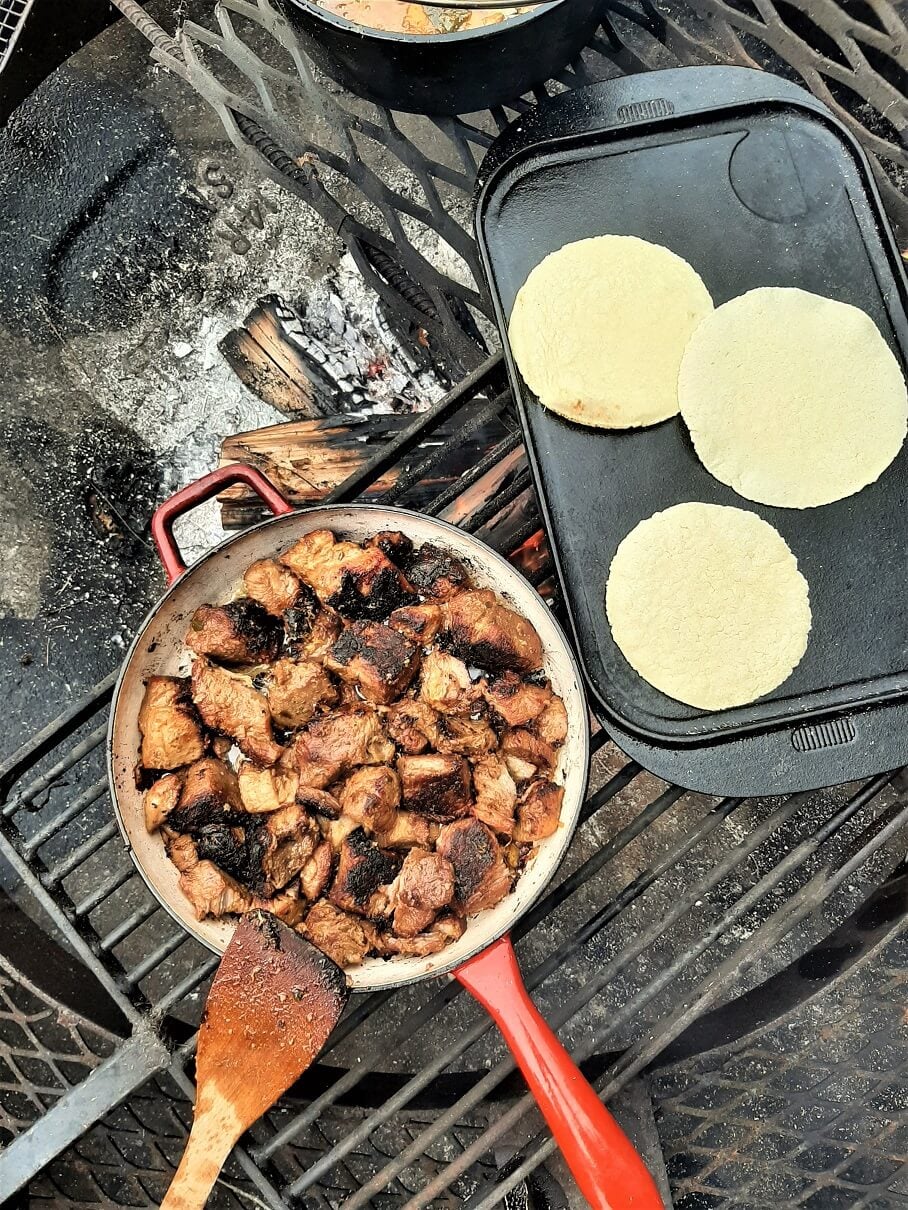 Close-up of two Instant Pot carnitas tacos on corn tortillas with crispy caramelized pork edges visible, topped with fresh cilantro, white onion, a squeeze of lime, and crumbled cotija cheese, with a blurred campfire and cast iron skillet in the background