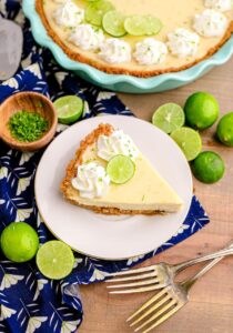 Classic Key Lime Pie with golden graham cracker crust sliced on a white cake stand with lime slices and whipped cream on top