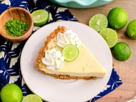 Classic Key Lime Pie with golden graham cracker crust sliced on a white cake stand with lime slices and whipped cream on top