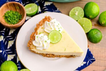 Classic Key Lime Pie with golden graham cracker crust sliced on a white cake stand with lime slices and whipped cream on top