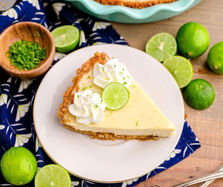 Classic Key Lime Pie with golden graham cracker crust sliced on a white cake stand with lime slices and whipped cream on top