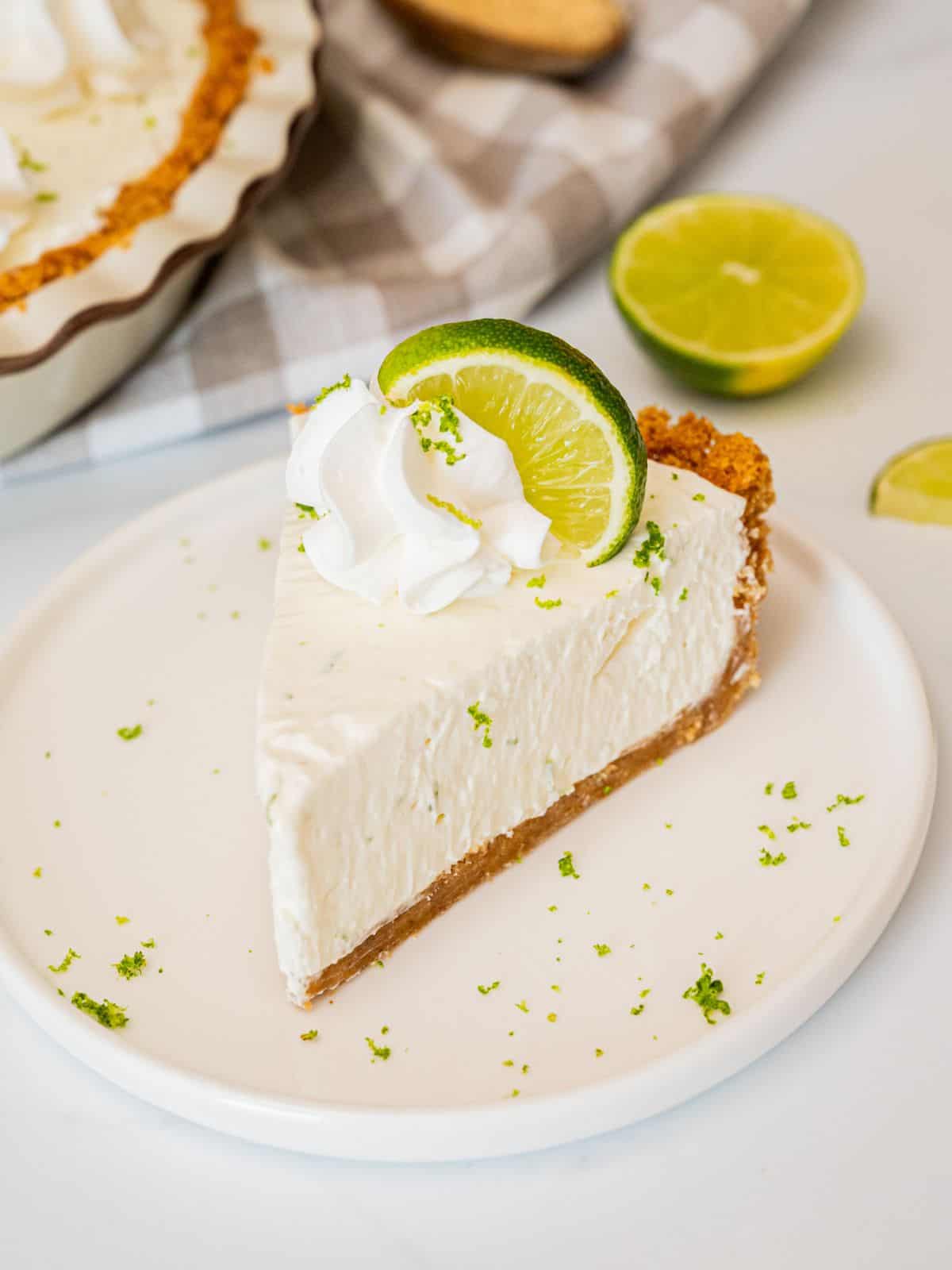 Close-up slice of Key Lime Pie on a white plate with swirl of whipped cream, lime zest, and thin lime wheel garnish on a wooden table
