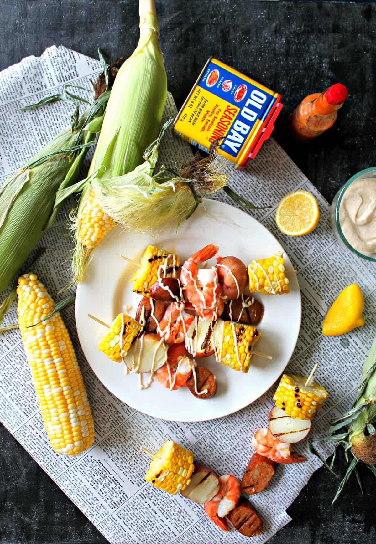 Overhead flat lay of Low Country Shrimp Boil ingredients including raw shrimp, corn cobs, baby potatoes, andouille sausage, lemon halves, garlic, and Old Bay seasoning on a white surface
