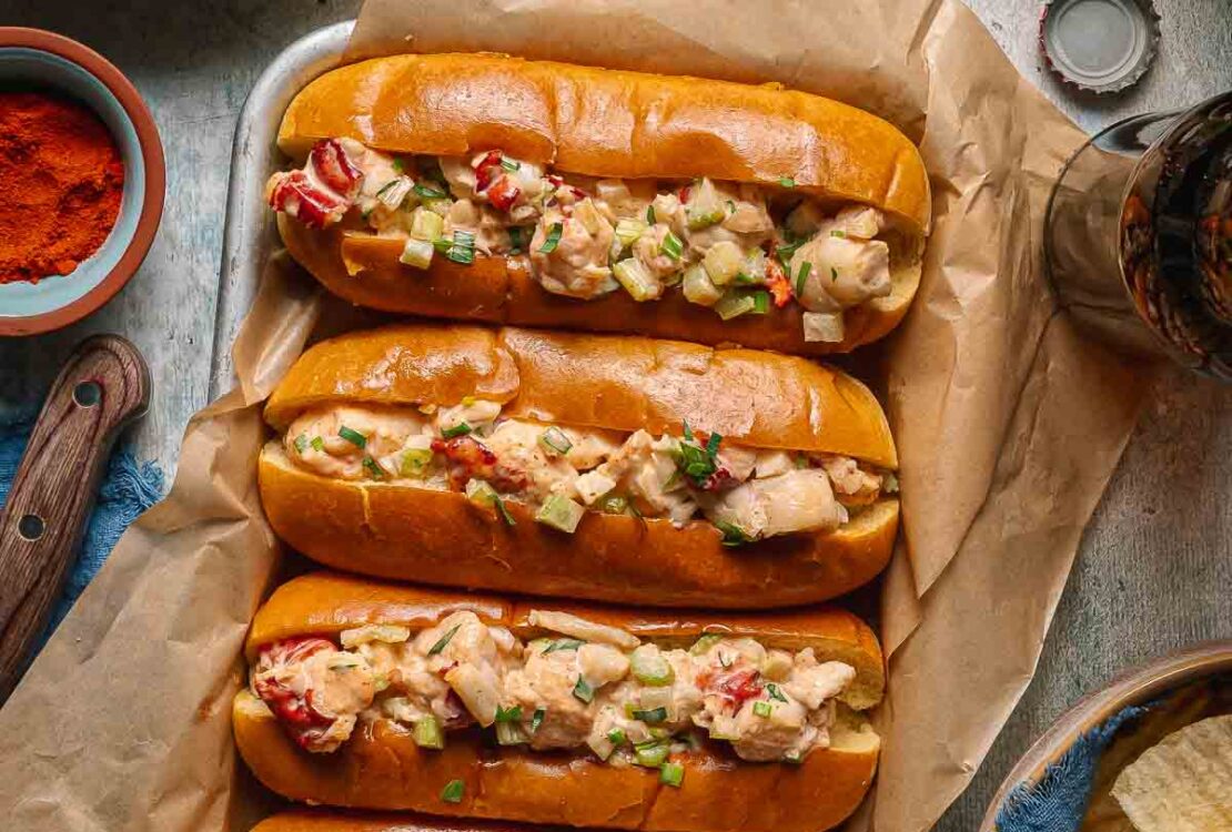 Classic Maine Lobster Rolls served in golden buttered split-top hot dog buns, piled high with creamy lobster salad, garnished with fresh chives and lemon wedges on a rustic wooden board