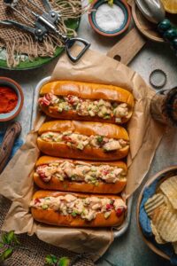 Classic Maine Lobster Rolls served in golden buttered split-top hot dog buns, piled high with creamy lobster salad, garnished with fresh chives and lemon wedges on a rustic wooden board