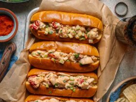 Classic Maine Lobster Rolls served in golden buttered split-top hot dog buns, piled high with creamy lobster salad, garnished with fresh chives and lemon wedges on a rustic wooden board