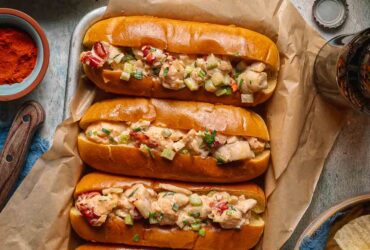 Classic Maine Lobster Rolls served in golden buttered split-top hot dog buns, piled high with creamy lobster salad, garnished with fresh chives and lemon wedges on a rustic wooden board