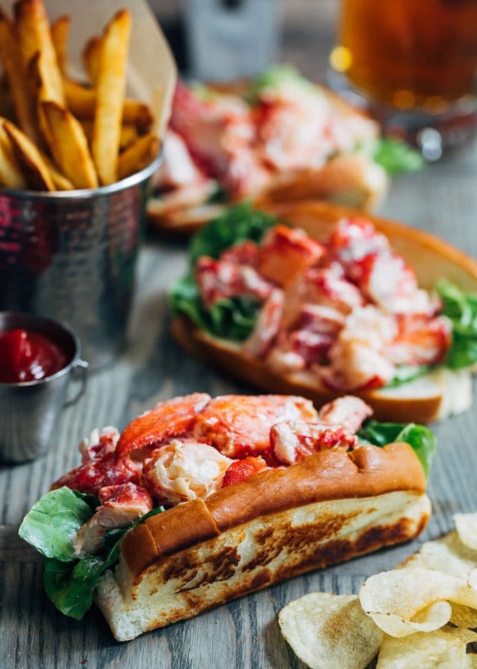Close-up of a Maine Lobster Roll split-top bun overflowing with creamy lobster salad, fresh chives, and a lemon wedge on the side, with a cold lemonade in the background on a sunny picnic table