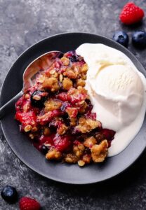 Mixed Berry Crisp with golden oat crumble topping served in a white baking dish with a scoop of vanilla ice cream melting over the top, styled on a rustic wooden table