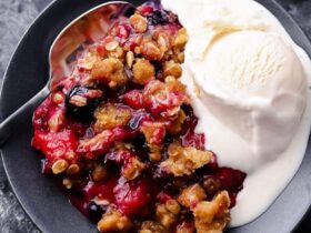 Mixed Berry Crisp with golden oat crumble topping served in a white baking dish with a scoop of vanilla ice cream melting over the top, styled on a rustic wooden table