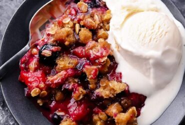 Mixed Berry Crisp with golden oat crumble topping served in a white baking dish with a scoop of vanilla ice cream melting over the top, styled on a rustic wooden table