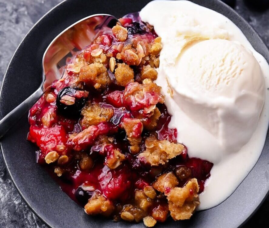 Mixed Berry Crisp with golden oat crumble topping served in a white baking dish with a scoop of vanilla ice cream melting over the top, styled on a rustic wooden table